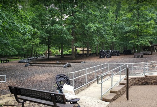 A park scene with benches, a stroller, and tire structures surrounded by trees and mulch.