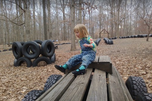 A child in overalls sits on a wooden platform in a forested area with tire structures and fallen leaves around.