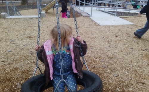 A young girl in a pink jacket swings on a tire swing at a playground, surrounded by wood chips and other children.