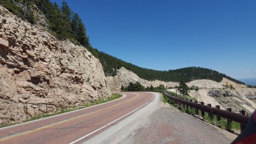 Winding road alongside rocky cliffs and green hills under a clear blue sky.