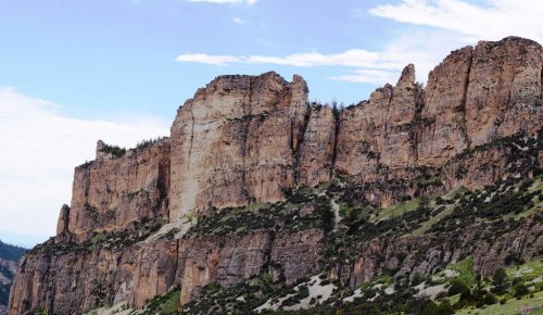 A rugged cliffside with steep rock formations under a blue sky, surrounded by greenery and distant mountains.