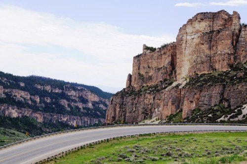 A winding road curves around rocky cliffs and green hills under a cloudy sky.