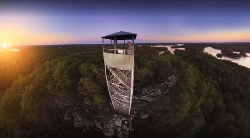 A tall observation tower stands on a rocky outcrop, surrounded by lush trees and a scenic sunset over a river.