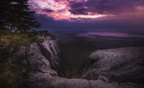 A rocky cliff overlooks a serene lake at sunset, with vibrant purple and pink clouds in the sky.