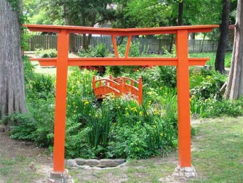 A vibrant orange torii gate stands in a lush garden, leading to a small red bridge over greenery and flowers.