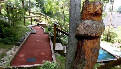 A wooden bear carving stands beside a mini-golf path surrounded by greenery and wooden structures.