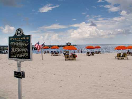 A beach scene with orange umbrellas, lounge chairs, and a historical marker about civil rights in the foreground.