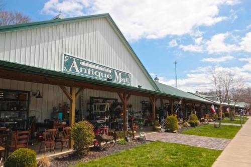 Exterior of an antique mall with a green roof, wooden beams, and various items displayed outside under a blue sky.