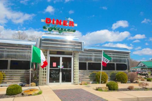 A retro diner with a shiny metal exterior, featuring flags of Italy and Mexico outside under a blue sky.