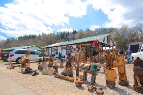 A rustic shop with wooden sculptures outside, surrounded by trees and a cloudy sky. Visitors browse the art.