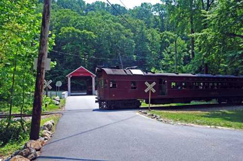 A vintage train passes a covered bridge, surrounded by lush green trees and a peaceful rural setting.