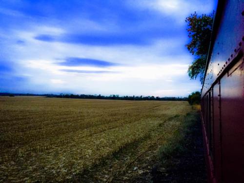 A train car alongside a vast, empty field under a cloudy blue sky.