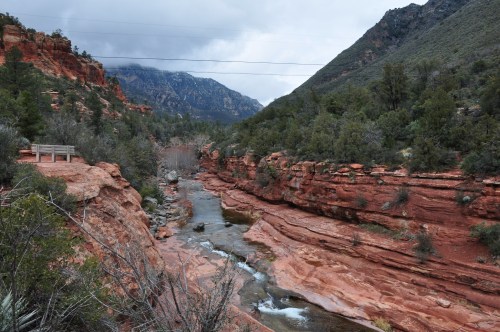 A scenic view of a winding river surrounded by red rock formations and green vegetation under a cloudy sky.
