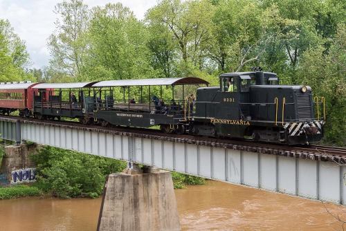 A vintage train crosses a bridge over a river, surrounded by lush green trees.