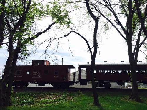 A red train car and a passenger car are silhouetted against a cloudy sky, surrounded by trees.