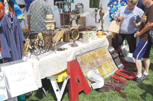 A bustling outdoor market stall displaying vintage items, mirrors, and decorative signs, with shoppers browsing.