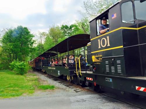 A black train with passengers on an open car travels through a green landscape under a cloudy sky.