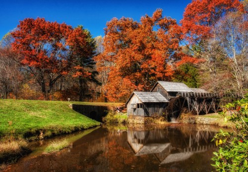 A scenic view of a wooden mill by a pond, surrounded by vibrant autumn foliage and clear blue skies.