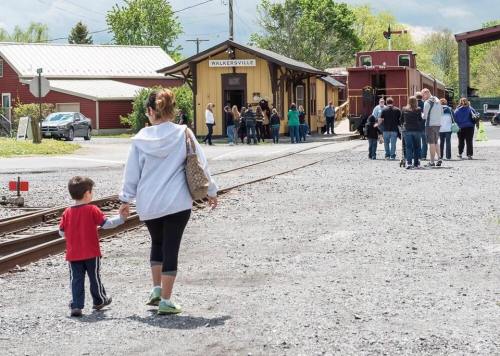 A woman and child walk hand in hand along a train track, with a crowd gathered near a station in the background.
