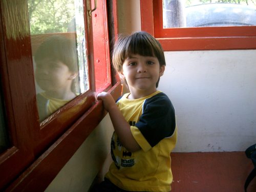 A young child in a yellow shirt sits by a window, smiling and looking at their reflection.