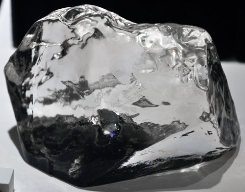 A large, clear, irregularly shaped crystal resembling ice, reflecting light and shadows on a surface.