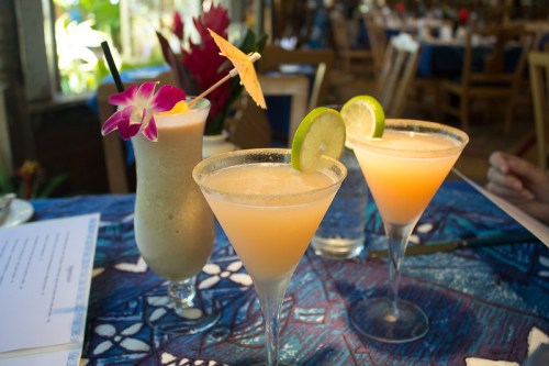 Three colorful cocktails on a vibrant table, garnished with fruit and umbrellas, set in a tropical dining atmosphere.