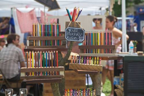 Colorful pencils arranged on wooden displays at an outdoor market, with a sign reading "Your Purchase Matters."