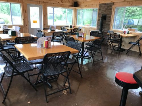 A cozy, empty dining area with wooden walls, tables, and chairs, featuring large windows and a stone fireplace.