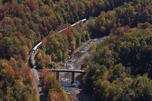 Aerial view of a train winding through colorful autumn trees near a river and a bridge.