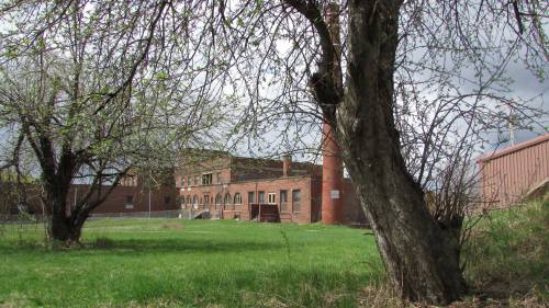 An old brick building with a chimney, surrounded by trees and grass under a cloudy sky.