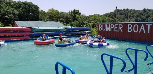 Four people in colorful bumper boats enjoy a sunny day on a water ride, surrounded by greenery and a sign that reads "BUMPER BOATS."