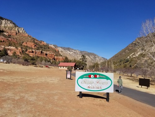 A scenic view of a market sign with mountains in the background and a person walking on a path. Clear blue sky.
