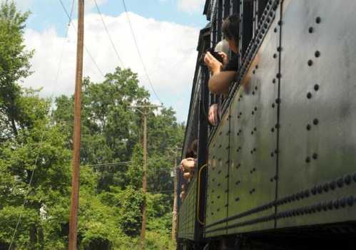 A vintage train passes by, with passengers leaning out the windows, surrounded by trees and power lines.