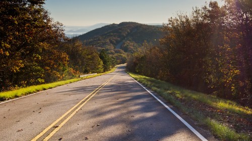 A winding road stretches through a scenic landscape of trees and hills under a clear blue sky.