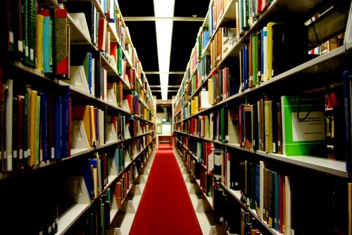 A narrow aisle between tall bookshelves filled with colorful books, with a red carpet running down the center.