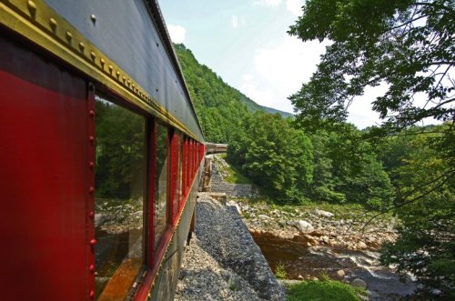 A scenic view of a train traveling alongside a river, surrounded by lush green trees and mountains.