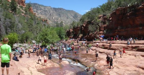 A crowded riverbank scene with people enjoying the water, surrounded by red rock formations and greenery under a clear blue sky.