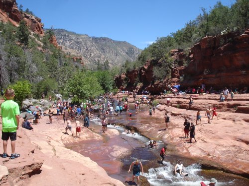A crowded river scene with people playing in the water, surrounded by red rock formations and greenery under a clear blue sky.