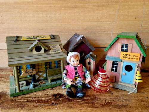 A wooden doll sits between two decorative birdhouses, one labeled "Hunting Lodge" and the other "Little Red School House."