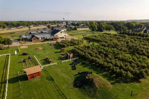 Aerial view of a farm with green fields, play structures, and an orchard, near a small town and highway.