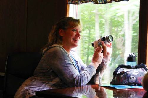 A woman smiles while holding a camera, sitting by a window with greenery visible outside.