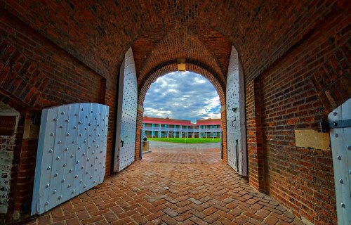 View through an arched doorway with large white doors, leading to a courtyard with a cloudy sky above.