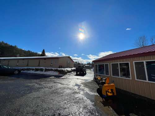 A clear blue sky with the sun shining over a parking lot, snow patches, and a building with a red roof.