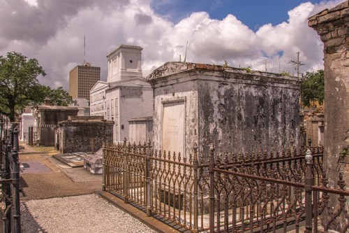 A historic cemetery with ornate tombs and mausoleums under a cloudy sky, surrounded by wrought iron fencing.