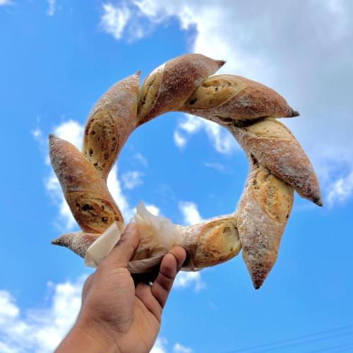 A hand holds a circular bread loaf against a blue sky with fluffy clouds.