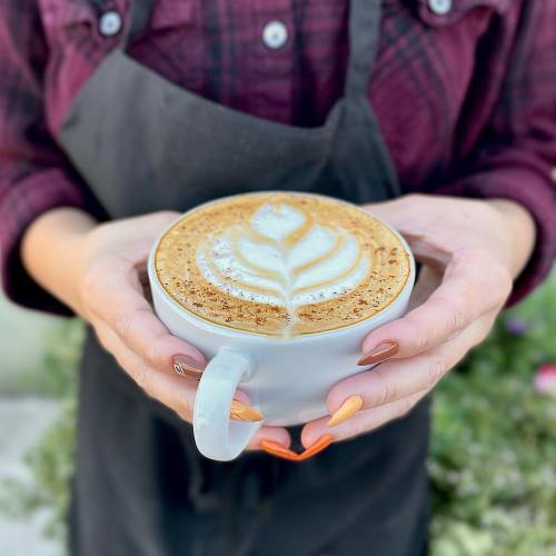 A person holding a white cup of latte art with a leaf design, wearing a plaid shirt and apron.