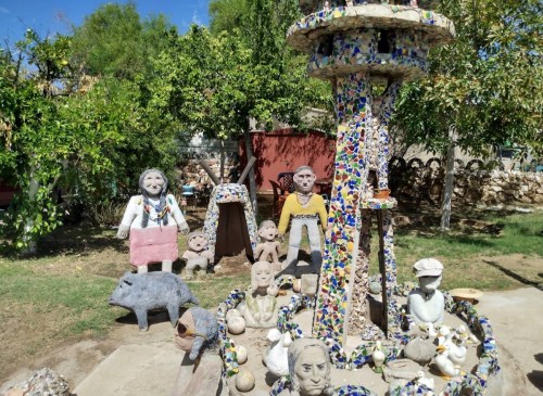 A whimsical outdoor scene featuring colorful sculptures of people and animals among greenery and a decorative fountain.