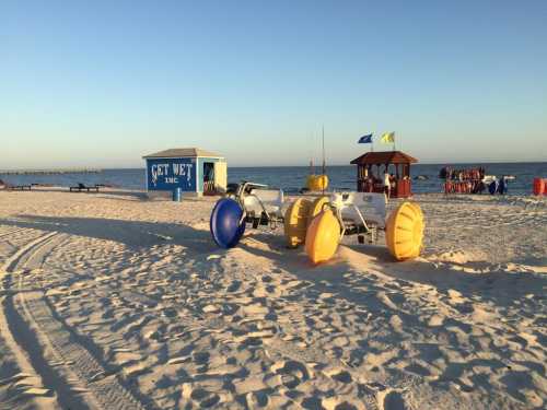 Beach scene with colorful watercraft on sand, a small building, and a pier in the background under a clear sky.