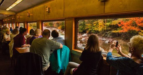 Passengers enjoy scenic views of a river and autumn foliage from a train window.