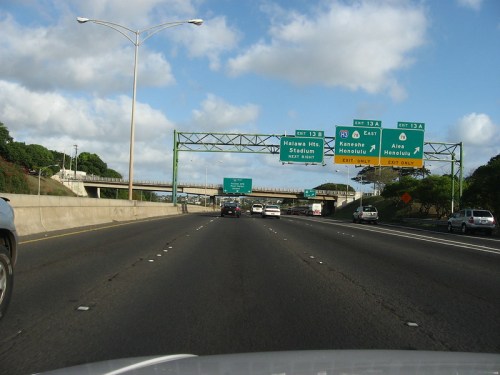 Highway scene with multiple exit signs for Halawe Hits Stadium and Honolulu, under a partly cloudy sky.